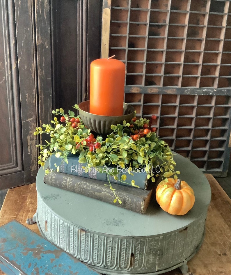 May include: A burnt orange pillar candle sits in a fluted metal holder atop two stacked vintage books. A faux greenery wreath with small red berries encircles the candle base. A small orange pumpkin rests nearby on a round, gray metal pedestal stand.