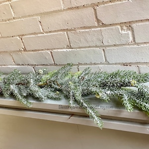 Peut inclure: Une guirlande artificielle de sapin enneig&eacute;e avec des aiguilles de pin vertes et des pointes givr&eacute;es, reposant sur un mur de briques blanches. La guirlande est parfaite pour ajouter une touche de joie hivernale &agrave; n'importe quel espace.