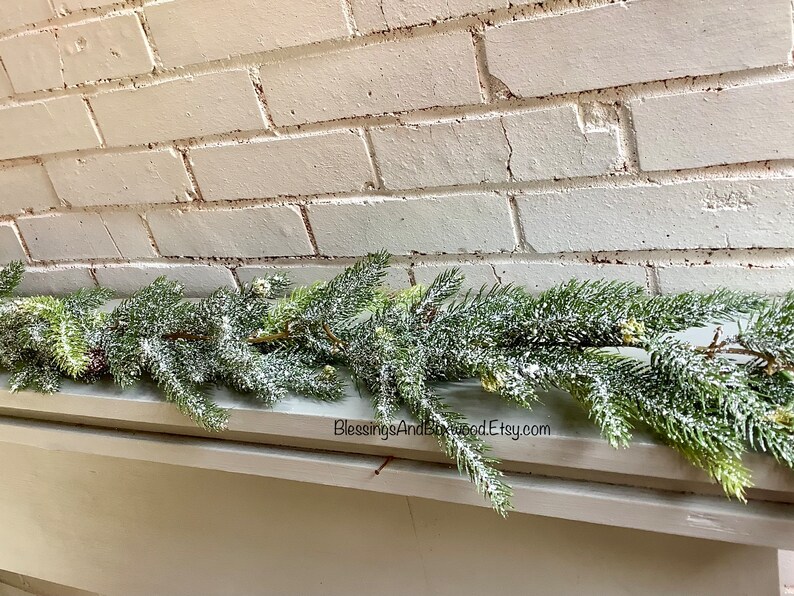 Peut inclure: Une guirlande artificielle de sapin enneig&eacute;e avec des aiguilles de pin vertes et des pointes givr&eacute;es, reposant sur un mur de briques blanches. La guirlande est parfaite pour ajouter une touche de joie hivernale &agrave; n'importe quel espace.