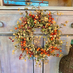 May include: A fall-themed wreath with orange berries and green and yellow leaves. The wreath is circular and hangs on a wooden cabinet. The text "Blessings And Boxwood Etsy" is visible on the wreath.