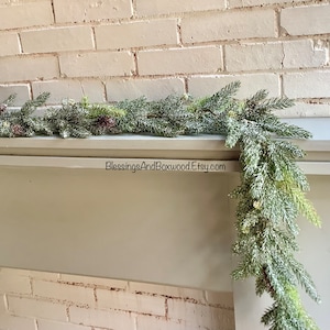 Peut inclure: Une guirlande de pin givr&eacute;e avec des baies rouges drap&eacute; sur une chemin&eacute;e blanche. La guirlande est d&eacute;cor&eacute;e de pommes de pin et a un aspect enneig&eacute; et hivernal. Le texte "BlessingsAndBoxwood.Etsy.com" est visible sur la chemin&eacute;e.