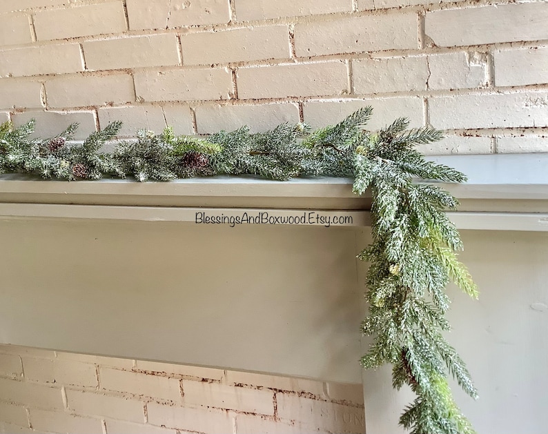 Peut inclure: Une guirlande de pin givr&eacute;e avec des baies rouges drap&eacute; sur une chemin&eacute;e blanche. La guirlande est d&eacute;cor&eacute;e de pommes de pin et a un aspect enneig&eacute; et hivernal. Le texte "BlessingsAndBoxwood.Etsy.com" est visible sur la chemin&eacute;e.