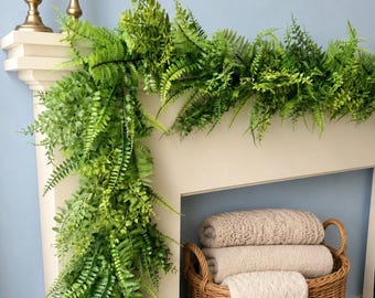 May include: A fireplace mantel decorated with a lush green fern garland. Two gold candle holders with white candles sit on the mantel. A woven basket holds folded blankets, creating a cozy, inviting scene.