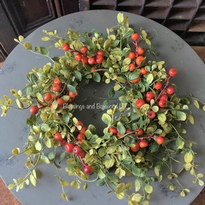 May include: A circular wreath featuring green boxwood leaves and clusters of small orange berries. The text Blessings and Boxwood LLC is printed in the center of the wreath. The foliage is arranged in a dense, rounded form with thin stems.