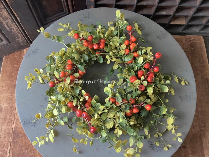 May include: A circular wreath featuring green boxwood leaves and clusters of small orange berries. The text Blessings and Boxwood LLC is printed in the center of the wreath. The foliage is arranged in a dense, rounded form with thin stems.