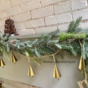 Peut inclure: Une guirlande de sapin enneig&eacute;e avec des clochettes dor&eacute;es est suspendue au-dessus d'une chemin&eacute;e. La guirlande est d&eacute;cor&eacute;e d'une pomme de pin et d'une corde de jute. Le texte "BlessingsAndBoxwoodEtsy.com" est visible sur la guirlande.