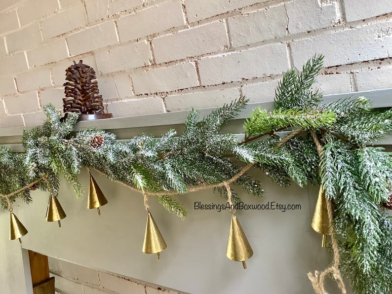 Peut inclure: Une guirlande de sapin enneig&eacute;e avec des clochettes dor&eacute;es est suspendue au-dessus d'une chemin&eacute;e. La guirlande est d&eacute;cor&eacute;e d'une pomme de pin et d'une corde de jute. Le texte "BlessingsAndBoxwoodEtsy.com" est visible sur la guirlande.