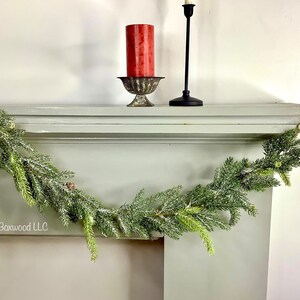 Peut inclure: Une guirlande floqu&eacute;e verte avec des aiguilles de pin et de petites pommes de pin est drap&eacute; sur une chemin&eacute;e peinte en blanc. Une bougie rouge est pos&eacute;e sur un bougeoir en m&eacute;tal et un bougeoir noir est sur la chemin&eacute;e.