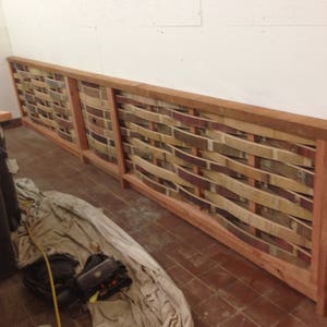 May include: A wall panel made of woven wood slats in shades of brown, beige, and red. The slats are arranged in a wavy pattern and are framed with a natural wood border.