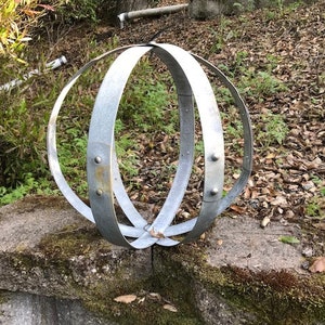 May include: A silver metal sphere made of three hoops, with a rustic, weathered finish. The sphere is sitting on a mossy stone wall.