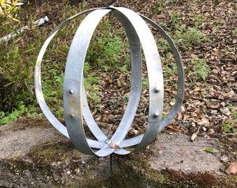 May include: A silver metal sphere made of three hoops, with a rustic, weathered finish. The sphere is sitting on a mossy stone wall.