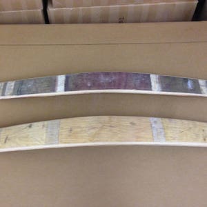 May include: Two wooden barrel stave wall decor pieces. The top piece is stained a dark brown with a red wine stain in the center. The bottom piece is a natural wood color. Both pieces are curved and have a rustic look.