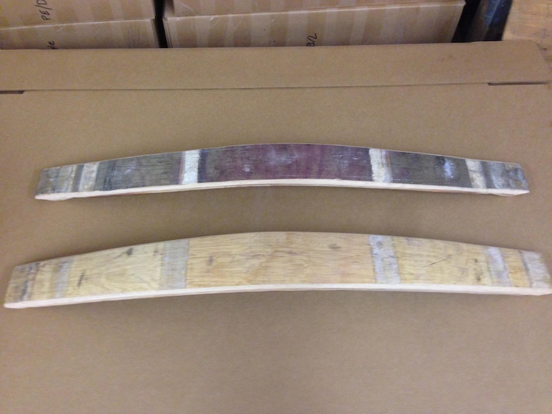 May include: Two wooden barrel stave wall decor pieces. The top piece is stained a dark brown with a red wine stain in the center. The bottom piece is a natural wood color. Both pieces are curved and have a rustic look.