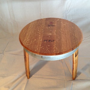 May include: A round wooden side table made from a repurposed barrel. The tabletop is a light brown color with visible wood grain. The table has three curved wooden legs and a metal band around the base of the tabletop. The text "GEM+T" is visible on the tabletop.