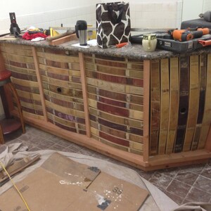 May include: A wooden bar counter with a curved front, constructed from reclaimed wine barrel staves. The counter is stained a light brown color and has a granite countertop. The counter is partially assembled and is sitting on a tiled floor.