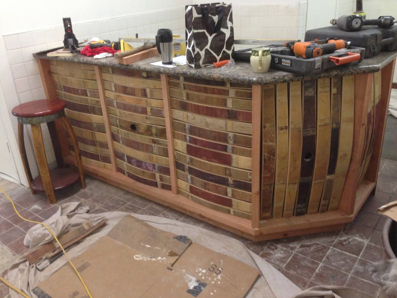 May include: A wooden bar counter with a curved front, constructed from reclaimed wine barrel staves. The counter is stained a light brown color and has a granite countertop. The counter is partially assembled and is sitting on a tiled floor.