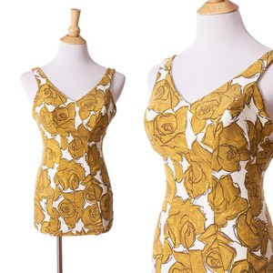 May include: A vintage-style dress with a floral pattern. The dress features a V-neckline and shoulder straps. The fabric is white with a gold rose pattern. The dress is displayed on a mannequin. The dress has a fitted bodice and a straight silhouette.