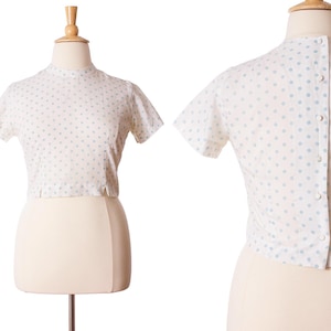 Puede incluir: Top vintage blanco de manga corta con cuello alto y lunares azul claro. La parte trasera del top presenta un cierre abotonado. El top se muestra en un maniquí.