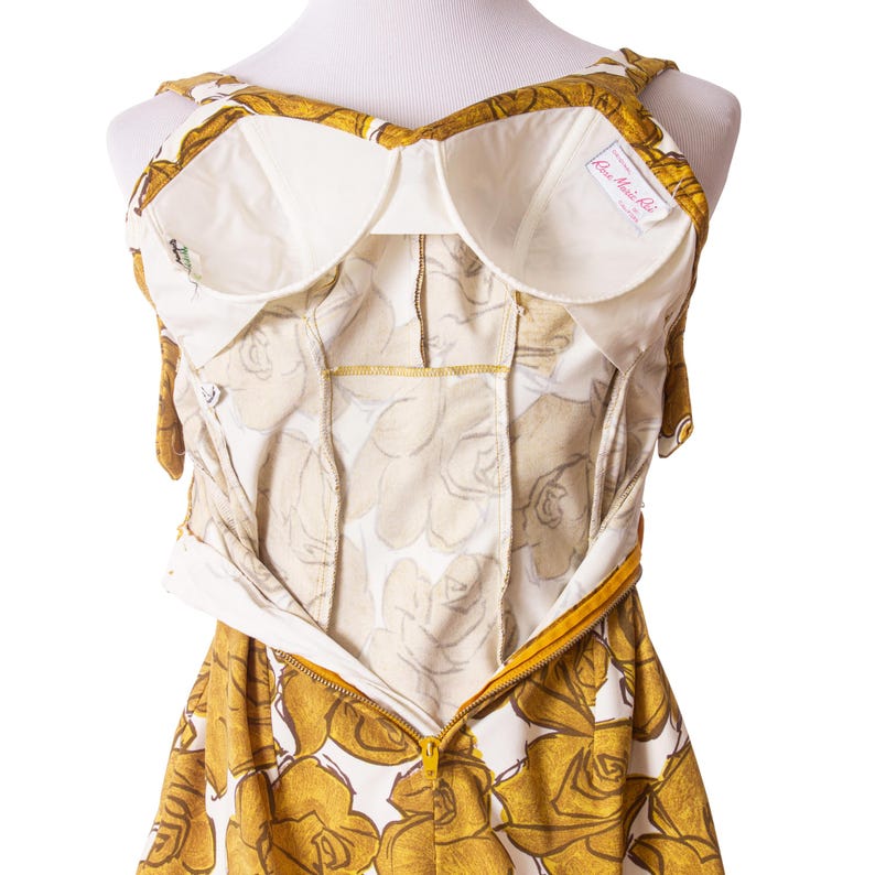 Op de afbeelding: Een vintage jurk met een bloemenpatroon in goud- en wittinten. De jurk heeft een ingebouwde bh met witte voering en een gele rits. De jurk wordt op een mannequin gepresenteerd.