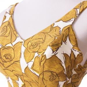 Op de afbeelding: Close-up van een mouwloze jurk met een bloemenpatroon. De jurk heeft een V-hals en dunne bandjes. Het bloemmotief toont grote, gestileerde rozen in goud- en bruintinten tegen een witte achtergrond.