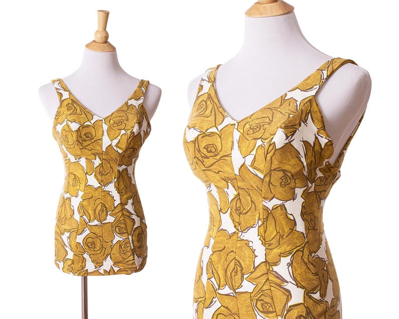 May include: A vintage-style dress with a floral pattern. The dress features a V-neckline and shoulder straps. The fabric is white with a gold rose pattern. The dress is displayed on a mannequin. The dress has a fitted bodice and a straight silhouette.