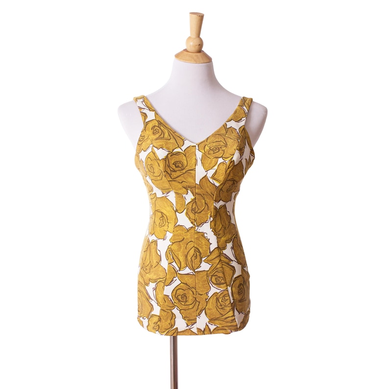 Op de afbeelding: Een vintage badpak met een V-hals, met een patroon van grote goudgele rozen op een witte achtergrond. Het badpak wordt op een mannequin getoond, waarbij het ontwerp en de pasvorm worden getoond. De bandjes zijn dun.