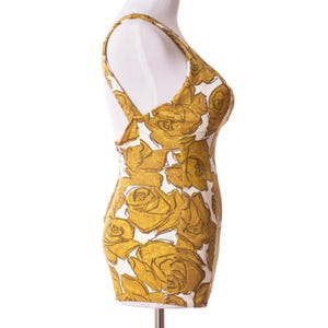 Op de afbeelding: Een vintage badpak met een bloemenpatroon. Het badpak heeft een witte achtergrond met grote, goudgele rozenillustraties. Het heeft een ronde halslijn en dunne schouderbandjes, wat een retro esthetiek laat zien.