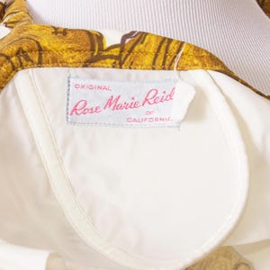 Op de afbeelding: Close-up van een vintage kledingstuk met een witte voering en een bloemenpatroon in goud- en bruintinten. Het label luidt "Rose Marie Reid of California."