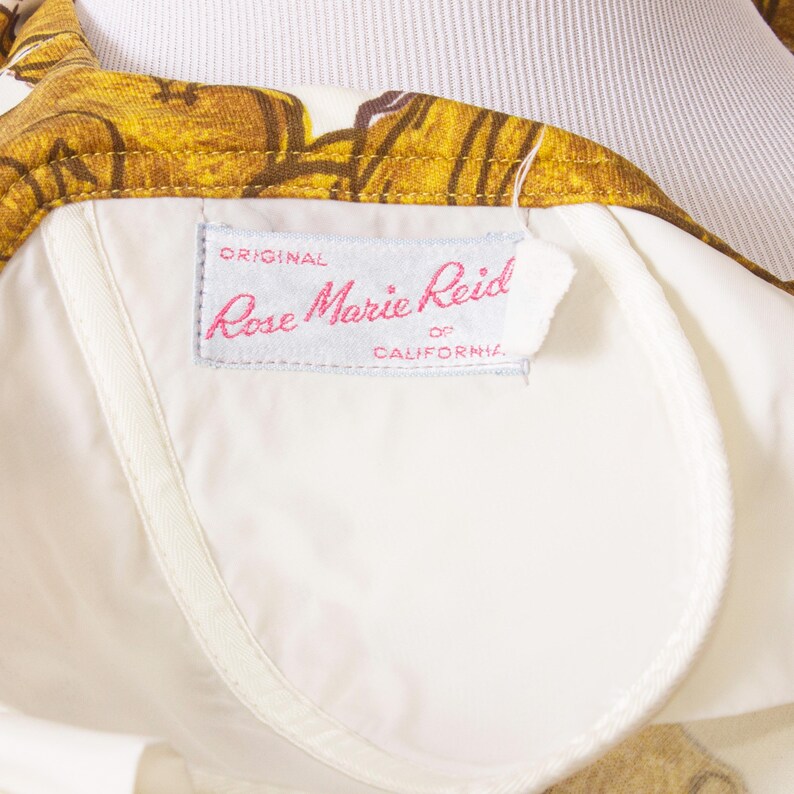 Op de afbeelding: Close-up van een vintage kledingstuk met een witte voering en een bloemenpatroon in goud- en bruintinten. Het label luidt "Rose Marie Reid of California."