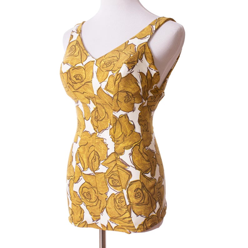 Op de afbeelding: Een vintage badpak met een V-hals en schouderbandjes. Het eendelige badpak heeft een witte achtergrond met een patroon van grote, goudgele rozen. Het kledingstuk wordt op een mannequin gepresenteerd tegen een witte achtergrond.