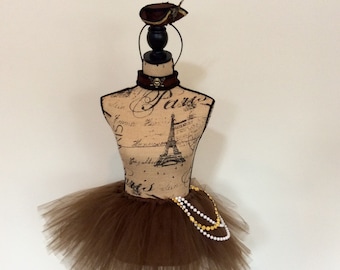May include: A brown tulle tutu skirt with a gold and white beaded necklace. The skirt is on a mannequin with a brown hat and a skull and crossbones choker.