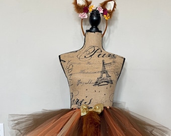 May include: A brown, orange, and beige tulle tutu with a fluffy brown and white fox tail attached to the back. A fox ear headband with pink and white flowers is included.