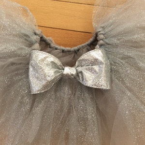 May include: A silver glitter tulle tutu skirt with a matching bow. The skirt is made of layers of shimmering tulle, gathered at the waist. The bow is made of a shiny silver material and is centered on the front of the skirt.