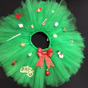 May include: A green tulle tutu with a red bow and various Christmas-themed decorations, including ornaments, candy canes, and a snowman. The tutu has the words "Merry Christmas" written in gold glitter.