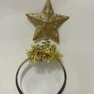 May include: A gold glitter star tree topper with a spiral base sits above a gold headband with gold tinsel. The headband is black on the inside. Festive holiday accessory.