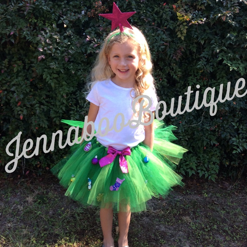 May include: A green tulle tutu skirt with a pink bow and various Christmas ornaments attached. The skirt is worn by a young girl wearing a white t-shirt and a red star headband. The girl is standing in front of a green bush.
