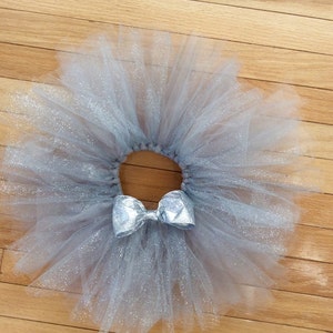 May include: A silver-gray tutu skirt with a matching bow. The skirt is made of layers of tulle, creating a full, fluffy appearance. The bow is centered on the waistband, adding a decorative touch. The tutu is designed for dance or dress-up.