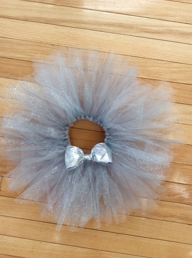 May include: A silver-gray tutu skirt with a matching bow. The skirt is made of layers of tulle, creating a full, fluffy appearance. The bow is centered on the waistband, adding a decorative touch. The tutu is designed for dance or dress-up.