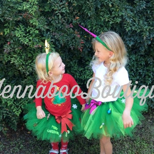 May include: Two girls wearing green Christmas tree tutus with festive decorations. One girl has a gold star headband and the other has a green and pink headband. The tutus are made of tulle and have a green and white color scheme. The girls are smiling and looking at each other.