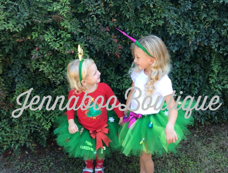 May include: Two girls wearing green Christmas tree tutus with festive decorations. One girl has a gold star headband and the other has a green and pink headband. The tutus are made of tulle and have a green and white color scheme. The girls are smiling and looking at each other.