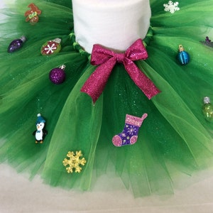 May include: A green tulle tutu with a pink bow and various Christmas ornaments, including a stocking, a penguin, a snowflake, and a light bulb.