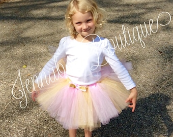 May include: A young girl wearing a white long-sleeved shirt and a pink and gold tulle tutu skirt with a gold bow. She is wearing pink shoes and standing on a gray surface.