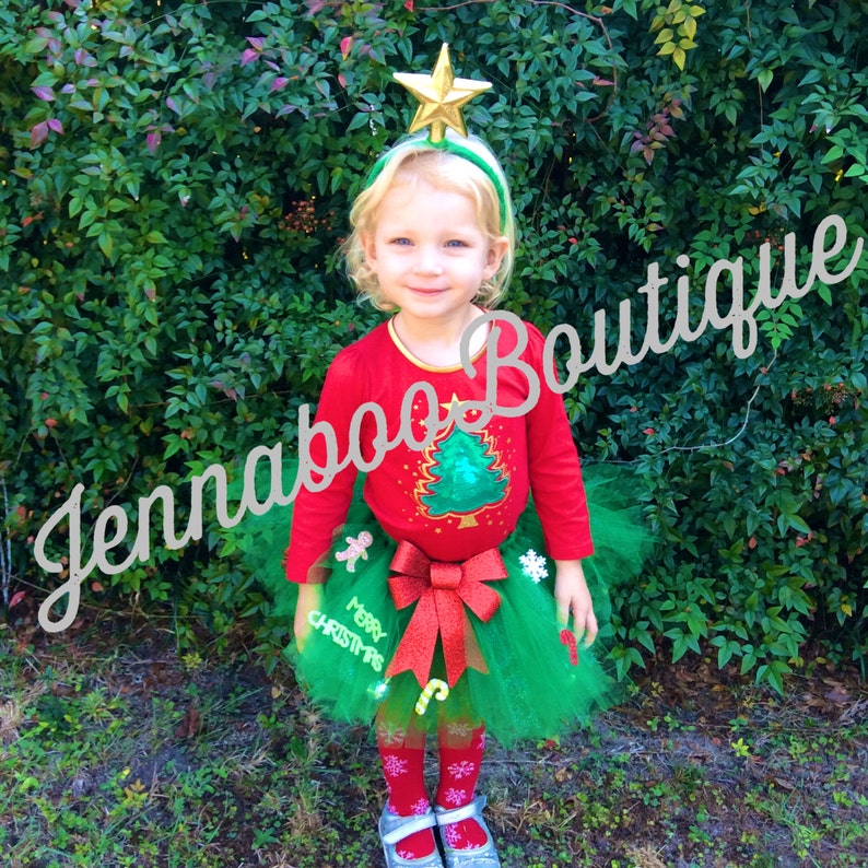 May include: A young child wearing a red Christmas tree shirt and a green tulle skirt with a red bow and Christmas decorations. The child is wearing a gold star headband and has a happy expression.