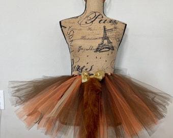 May include: A brown, orange, and beige tulle tutu with a fluffy brown tail and a gold bow. The tutu is on a mannequin wearing a brown headband with faux fur ears and pink and burgundy flowers.