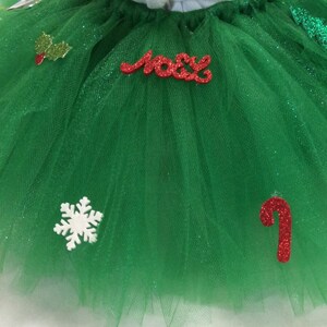 May include: Green tulle tutu with red and white glitter embellishments, including a snowflake, a candy cane, holly leaves, and the word "Noel".