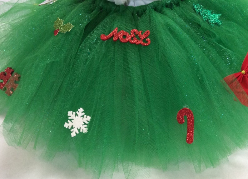 May include: Green tulle tutu with red and white glitter embellishments, including a snowflake, a candy cane, holly leaves, and the word "Noel".
