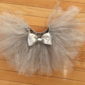 May include: A silver glitter tulle tutu skirt with a matching silver bow. The skirt is made of layers of soft, shimmering tulle and is designed for children. The tutu is a light gray color.