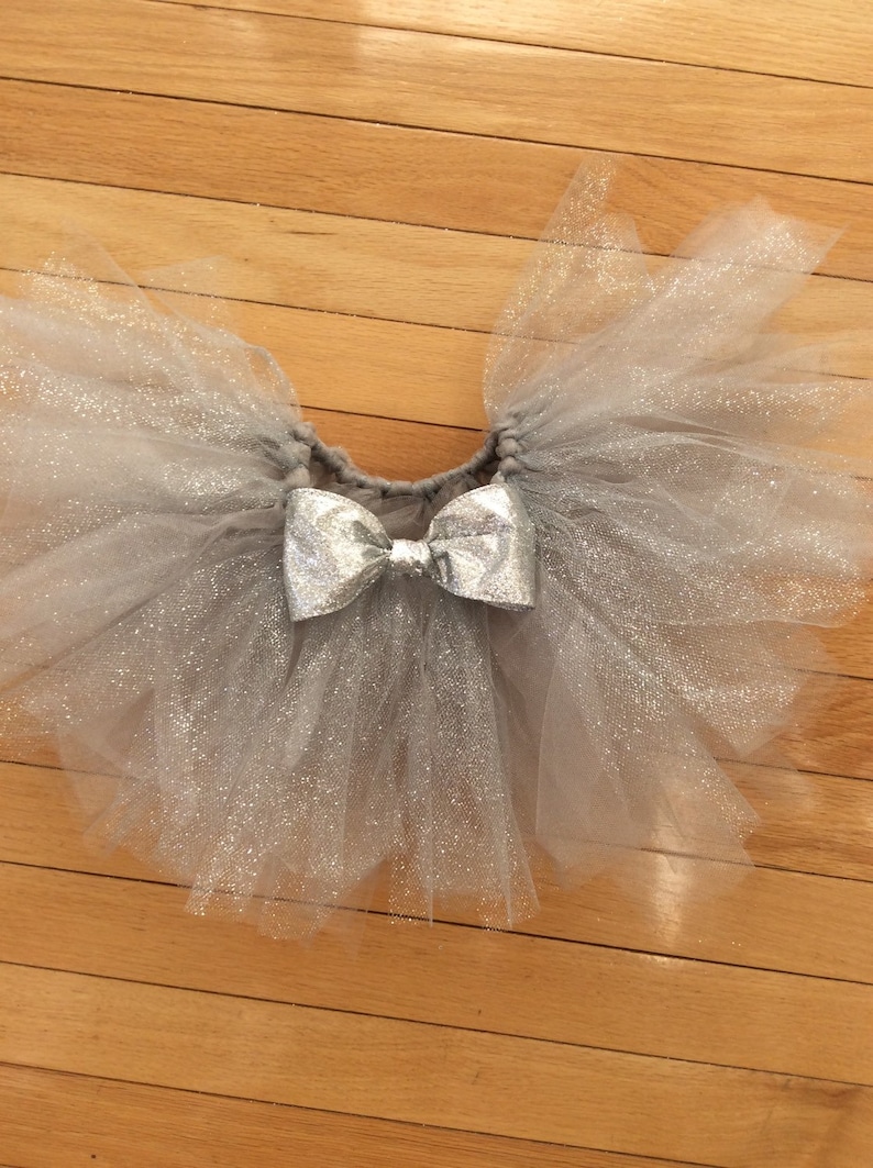 May include: A silver glitter tulle tutu skirt with a matching silver bow. The skirt is made of layers of soft, shimmering tulle and is designed for children. The tutu is a light gray color.