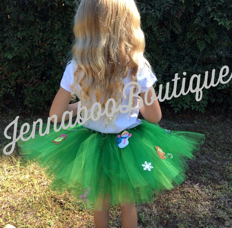 May include: A green tulle tutu skirt with various Christmas-themed felt appliques, including a snowman, a snowflake, and a gingerbread man. The skirt is worn by a young girl with long blonde hair. The text "Jennaboo Boutique" is visible in the background.