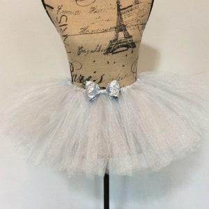 May include: A light blue tutu skirt with a silver bow. The skirt is made of layers of tulle and is displayed on a mannequin. The bow is centered on the waistband.